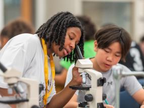 A student looking into a microscope while another student takes notes