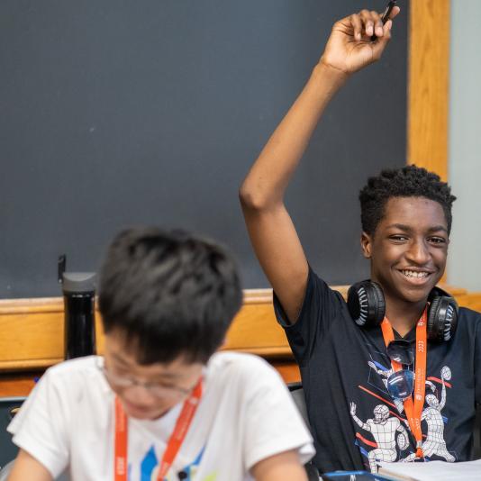 A student sitting at a desk, smiling, and raising his hand.