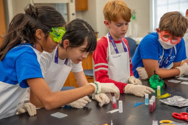 Four young advanced learners wearing lab goggles and aprons while conducting a science experiment.