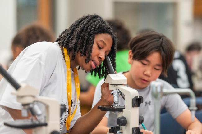 A student looking into a microscope while another student takes notes